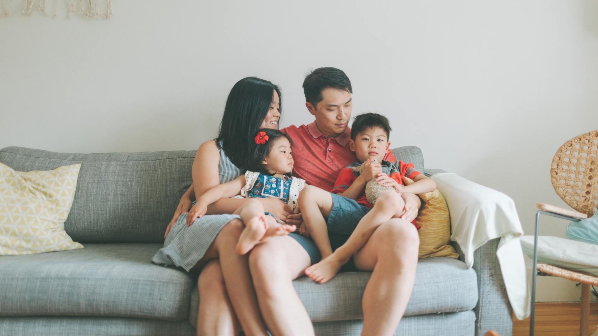 Asian american couple sitting on grey couch each with a child on their lap approximately 4-6 years of age