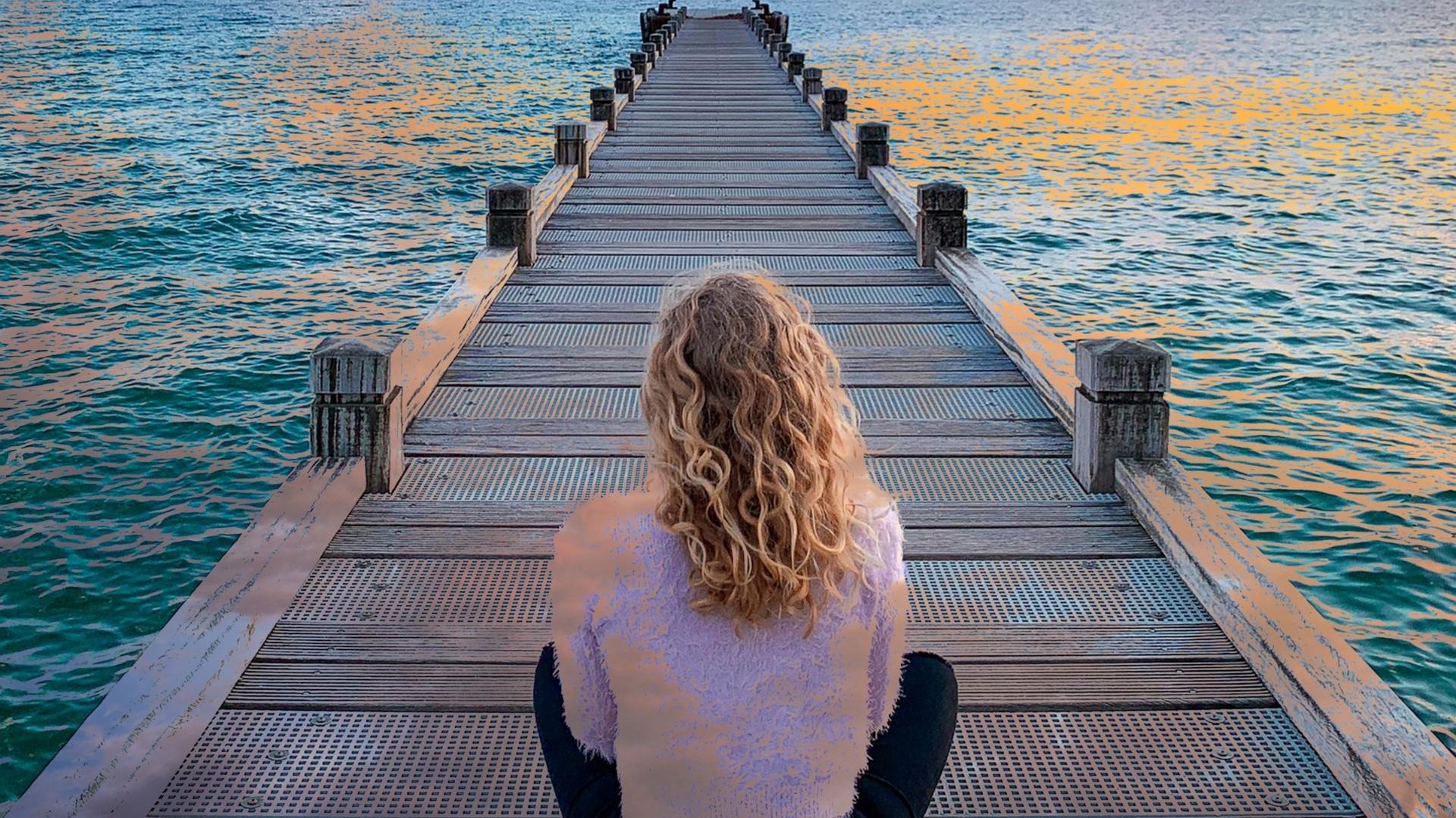 Woman sitting on long dock over blue and organe sunset reflection in water