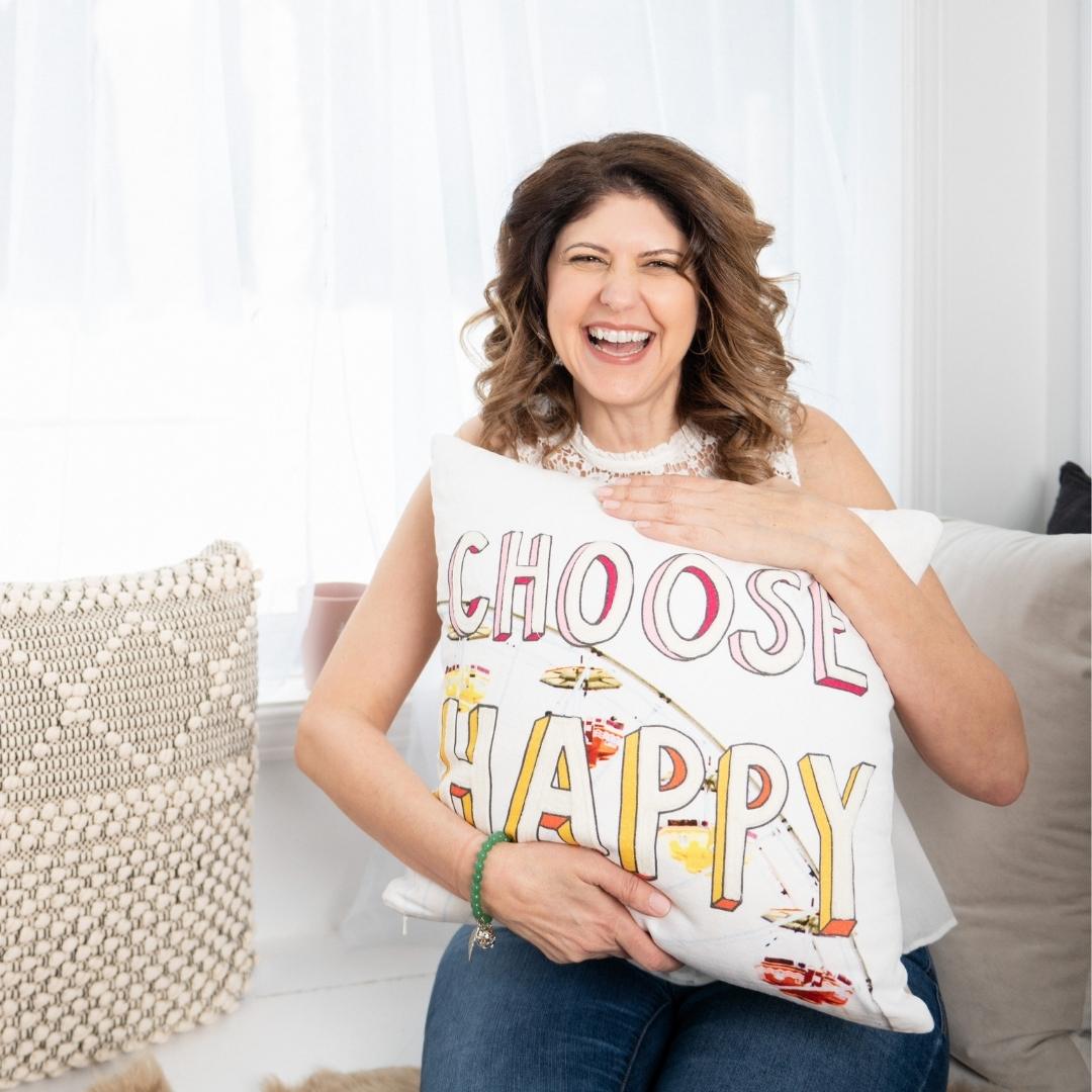 Marie, middle-aged white woman white shoulder length brown hair. holding pillow that says Choose Happy. White sheer curtains.