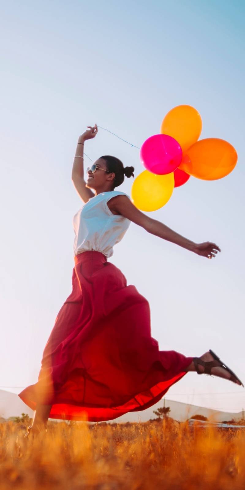 Woman with 5 balloons in orange pink and red. Hoping in a field. Red long skirt. White tank top. Blue Sky. 